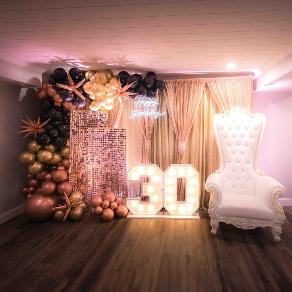 A custom party setup for someone's 30th birthday party. A white Victorian style chair, with multi-colored balloons with peach colored tapestry.